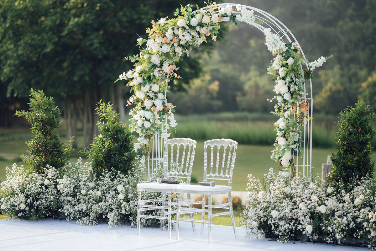 A beautifully decorated outdoor wedding arch with white chairs. The arch is adorned with flowers and greenery.