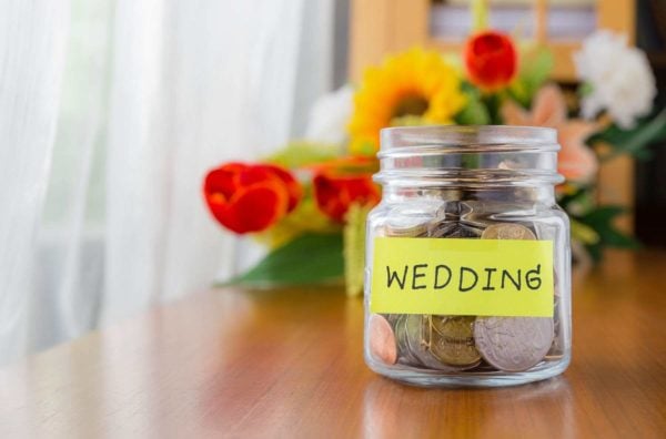 A glass jar labeled 'Wedding' filled with coins, sitting on a table with a bouquet of colorful flowers in the background.