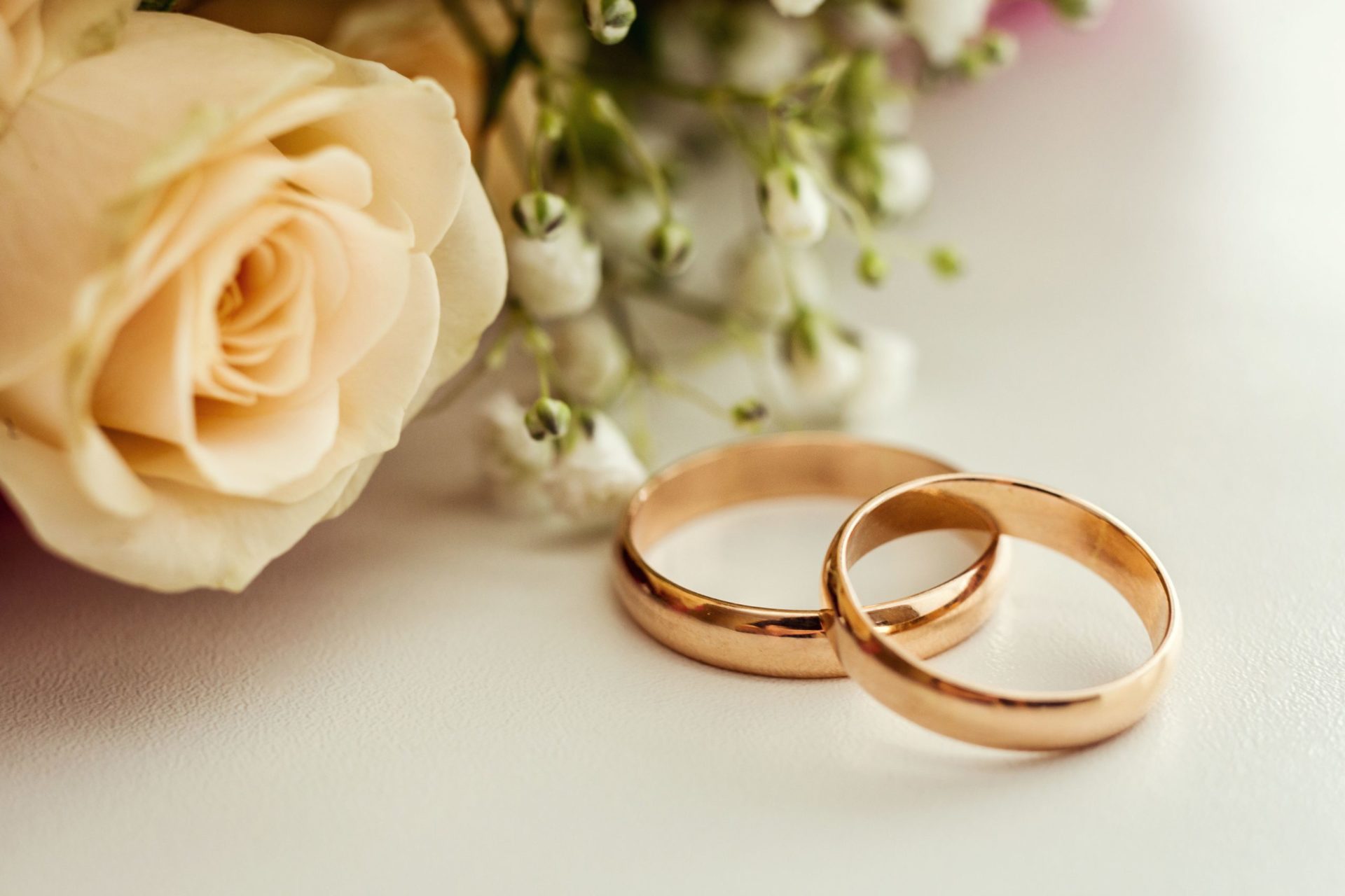 Two gold wedding bands on a white surface with a rose and baby's breath flowers in the background. The image symbolizes love and commitment.