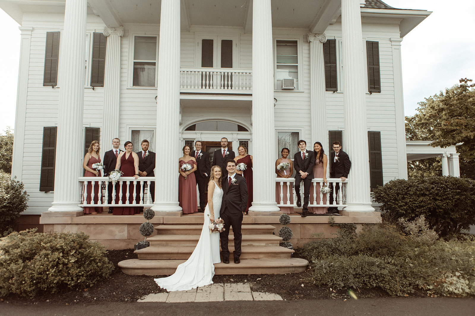 Bride and groom standing on steps with wedding party. The bride wears a white dress and the groom is in a black suit.