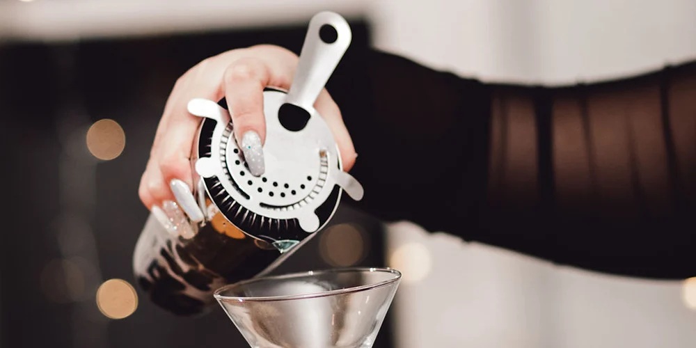 A bartender's hand holding a strainer and pouring a cocktail. The drink is being strained into a martini glass.
