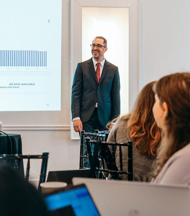 Businessman giving presentation to audience A man in a suit stands before a projector screen. People sit at desks in front of him.