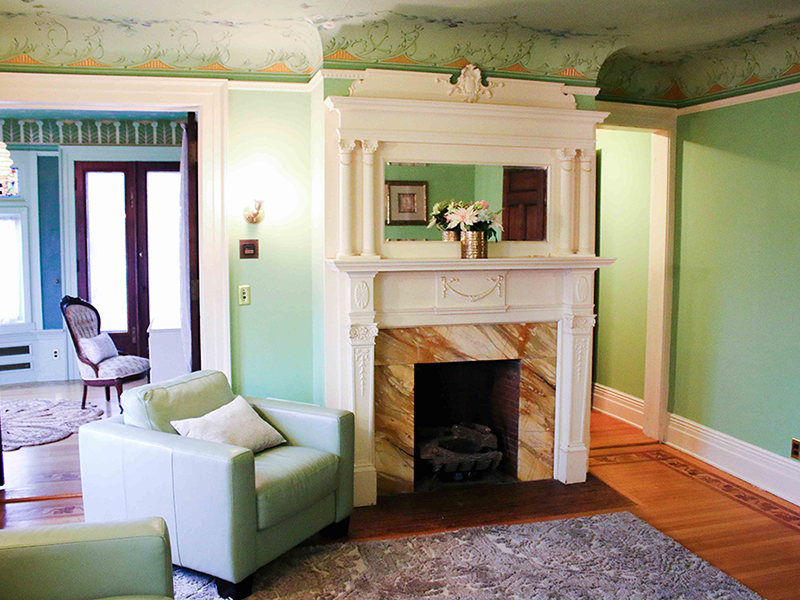 A vintage living room with a white fireplace and a green sofa. The room features a brown rug and wooden floor.