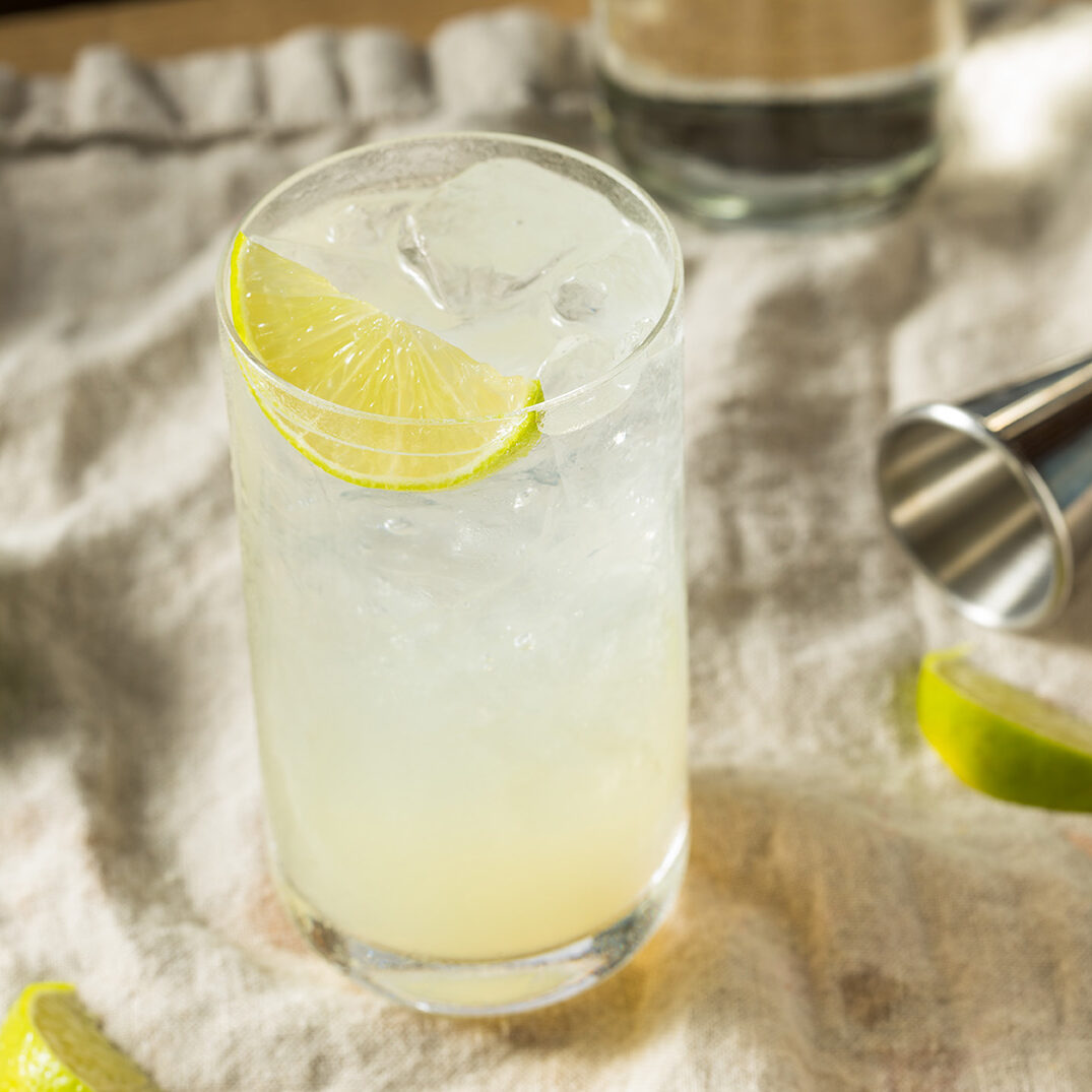 A glass of pale yellow cocktail with ice and a lime wedge, accompanied by a jigger and lime pieces on a beige cloth.
